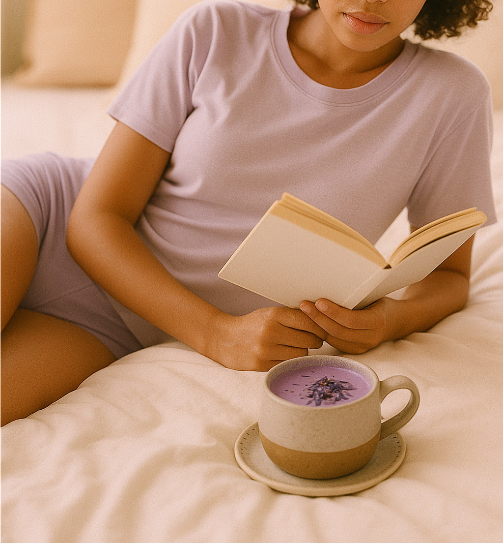 Woman in lavender loungewear reading a book in bed with a cup of Dreamy Soothe lavender taro latte on a saucer